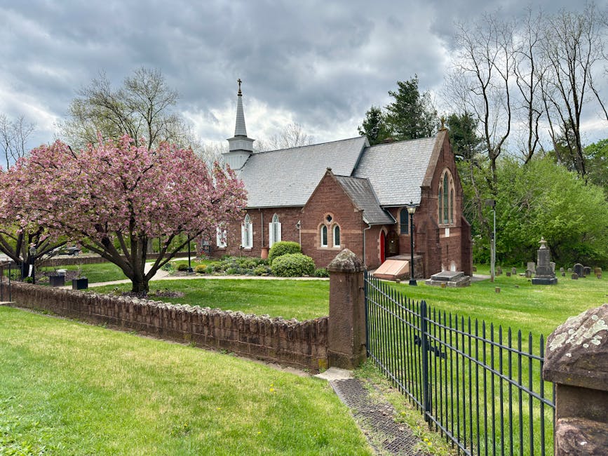 A vibrant pink flowering tree with full blossoms stands in front of a traditional red brick church with tall arched stained glass windows and a white spire, surrounded by a well-maintained grassy lawn