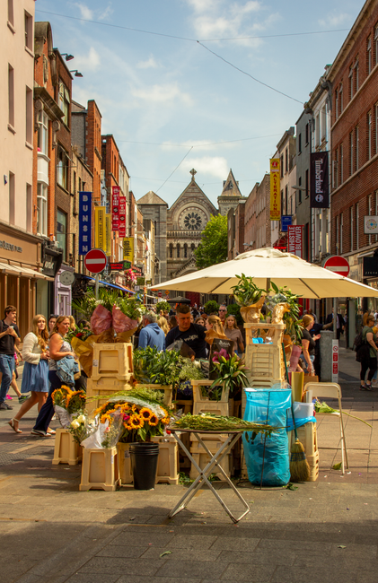 A vibrant street scene near Old Street in St Lukes, featuring an outdoor flower market with a variety of fresh floral arrangements and potted plants displayed on wooden crates and tables. The flowers 