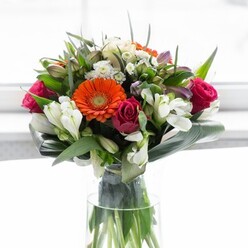 This magnificent bouquet, crafted with devotion in the heart of St Luke's, brings together the drama and delicacy of nature's finest blooms in an artful display. Creamy peach and fiery orange gerbera daisies form an inviting focal point, their soft petals unfolding around dark, textured centers that gleam under the gentle flow of natural light-much like the vibrant flower stalls at Whitecross Market. Deep fuchsia roses, their edges kissed with romance, nestle among the gerberas, imparting a sense of luxury and heartfelt affection. Sprinkles of snow-white alstroemeria and small, star-shaped filler blooms peek through the greenery, illuminating the bouquet with a bright, garden-fresh feeling. The entire composition is embraced by large, glossy aspidistra leaves expertly curved to create a rich, protective collar-a detail reflecting the quiet strength and warmth of the St Luke's community. Between these, pointed emerald leaves and fine green sprigs add subtle contrast, while the clear glass vase showcases the lively green stems, evoking freshness sourced from the city's best morning markets. The soft illumination against a pale backdrop makes every hue glow, suggesting renewal and gentle celebration. This bouquet, evocative of new beginnings, warm gatherings, or thoughtful appreciation for someone special in St Luke's, carries a subtle, rejuvenating fragrance and a whisper of hope with every bloom.